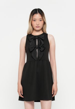 Robe noire avec un décolleté rond, sans manches, et un accent de nœud proéminent au niveau de la poitrine. Silhouette fluide avec un tissu texturé.