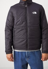 Chaqueta acolchada negra aislante con cuello alto, cierre de cremallera y logotipo en el lado izquierdo del pecho. Presenta un diseño acolchado y puños elásticos.