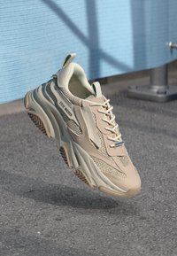 Beige sneaker with mesh and synthetic upper, lace-up design, thick sole, notable branding on the side, and textured rubber outsole.