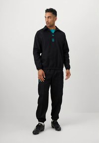 Fekete atlétikai melegítő gallérral, türkizkék kiemelésekkel a mellkason és a nadrágon. Az anyag simának tűnik; testhezálló szabás és rugalmas mandzsetták.