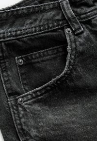 Gros plan sur une poche avant de jean en denim noir avec des coutures visibles, une passants de ceinture et des détails en rivets métalliques.