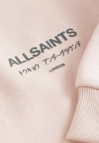 Ljusrosa stickad tyg med svart text "ALLSAINTS," japanska tecken och "LONDON," som visar en veckad manschett på höger sida.