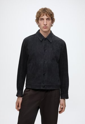 Göndör hajú férfi, fekete gombos nubuk dzsekit és sötét nadrágot visel, egyszínű fehér háttér előtt áll.