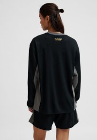 Zwarte sporttop met lange mouwen en ribbelstructuur, grijze accenten en een gouden logo op de rug, gecombineerd met zwarte shorts.