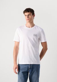 Ung mann med kort krøllete hår har på seg hvit Hugo-logo t-skjorte og blåjeans, står mot en enkel lys bakgrunn.