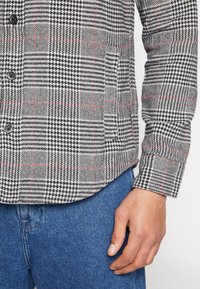 Man i svart och vitt houndstooth rutigt skjorta med röda linjer och blå jeans, med handen vilande vid sidan.