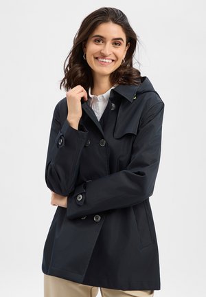 Smiling woman with shoulder-length dark hair wearing a black double-breasted coat over white shirt, posing with crossed arms on light background.