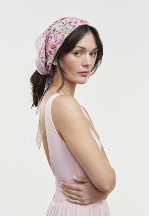 Mujer con el pelo oscuro recogido, lleva un pañuelo rosa con flores y un vestido sin mangas de color rosa pálido, mirando por encima del hombro con los brazos cruzados.