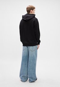 Μαύρη κουκούλα με μεγάλη κουκούλα, σε συνδυασμό με φαρδιά ανοιχτό μπλε τζιν παντελόνια που διαθέτουν πίσω τσέπες και έχει χαλαρή εφαρμογή.