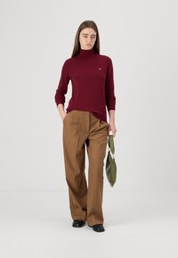 Burgunderfarget turtleneck-genser i myk strikk, sammen med brune høyt liv-bukser. Modellen holder en grønn håndveske og har på seg svarte sko.