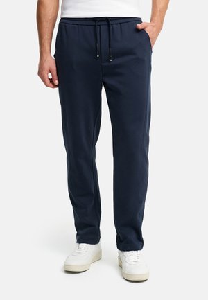 Homme portant un pantalon de survêtement bleu marine à cordon de serrage et des baskets blanches, debout avec une main dans la poche.