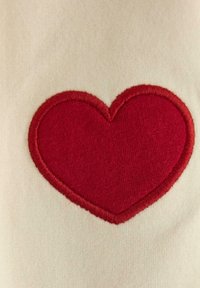 Beige stof med et stort rødt broderet hjerte. Hjertet har en struktureret overflade med en syet kant, der står i kontrast til den glatte baggrund.