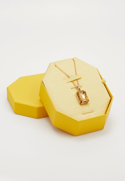 Collier en chaîne dorée avec un grand pendentif en ambre rectangulaire dans une boîte à bijoux octogonale jaune avec couvercle à côté sur fond blanc.