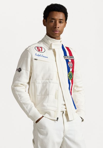 White quilted jacket with high collar, red and blue stripes, and multiple patches. Features a zip pocket and ribbed cuffs.