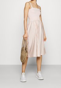 Axellös, smockad klänning i vitt med rosa vertikala ränder. Har en utsvängd kjol och ett lätt, texturerat tyg. Inkluderar en vävd stråväska.