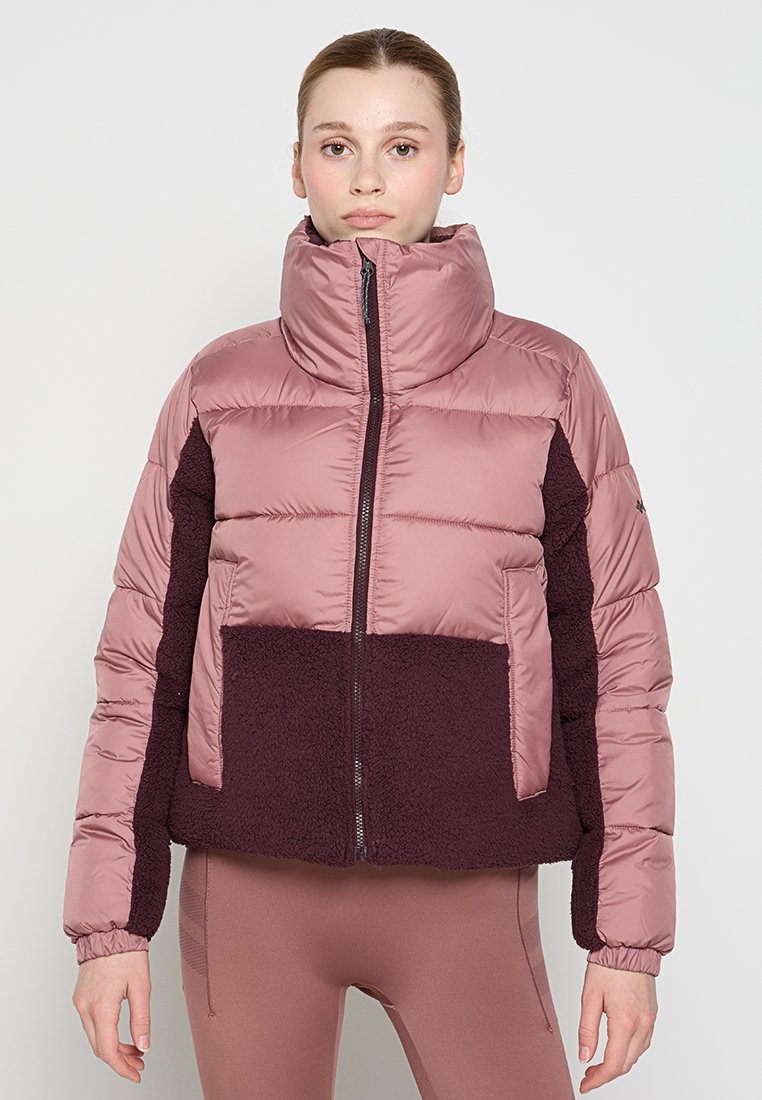 Frau, die eine zweifarbige, mauve und dunkelviolette Daunenjacke mit Fleece-Elementen trägt und vor einem schlichten weißen Hintergrund steht.