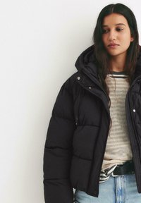 Zwarte gewatteerde jas met een capuchon, voorzien van een gekleed ontwerp en trekkoorddetails. Draagt over een gestreepte gebreide top en blauwe jeans.