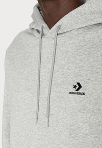 Sudadera con capucha de color gris claro que incluye una capucha con cordón, textura suave y logo bordado en negro de "Converse" con diseño de estrella y flecha.