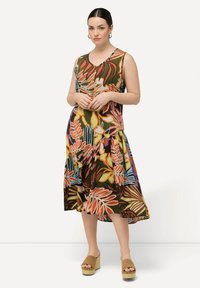 Vestido midi floral en tonos multicolores con base verde, que presenta un escote redondo y un dobladillo en capas. Combinado con sandalias de plataforma.