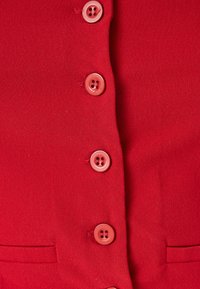 Rotes Stoffoberteil mit einem Knopf-Design, das sechs rosa Knöpfe umfasst, strukturierten Oberfläche und einem kleinen Taschen Detail auf der linken Seite.