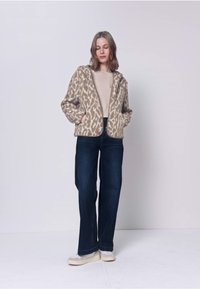 Frau trägt eine beige Jacke mit Leopardenmuster, ein hellbeiges Oberteil, dunkelblaue Jeans mit weitem Bein und beige Sneaker, steht vor einem schlichten hellen Hintergrund.