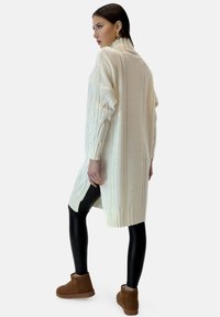 Strikket cremefarvet cardigan med høj krave, sideslids og ribbet kant, kombineret med sorte leggings og brune ankelstøvler. Glat tekstur.