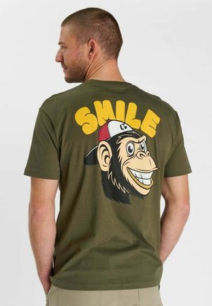 Uomo rivolto verso sinistra che indossa una maglietta verde oliva con una caricatura di uno scimpanzé sorridente con un cappello rosso e bianco e la parola "SMILE" in grandi lettere gialle.