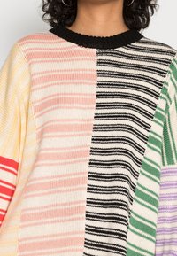 Strikket sweater med striber og en rund sort krave. Har sektioner i pastelrosa, gul, sort, grøn og lilla striber i forskellige størrelser.