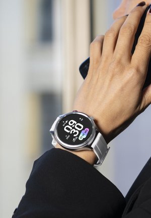 Håndled med et hvidt smartwatch, der viser klokken 09:30, datoen 28.08 onsdag og fitnessdata; hånd holder en sort smartphone tæt på øret.