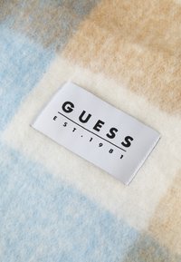 Flannel stof med blå, beige og hvide tern; et hvidt vævet mærke med mærket "GUESS" og "EST. 1981" i sort.