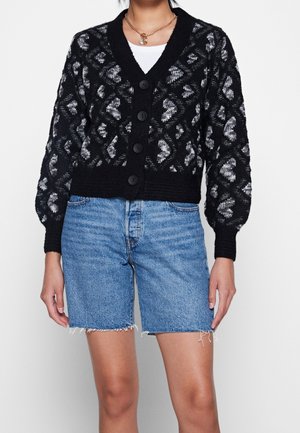 Femme portant un cardigan noir et gris à motifs avec de gros boutons sur un top blanc et un short en jean bleu clair aux bords effilochés.