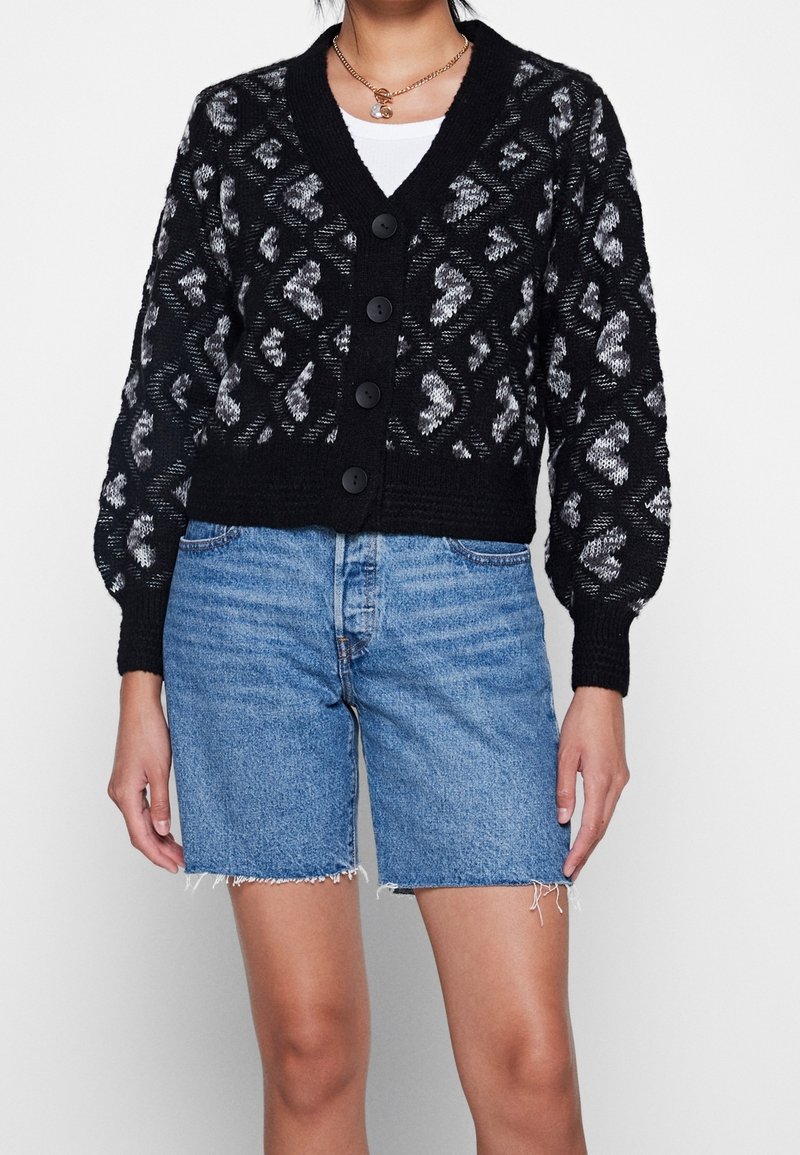 Femme portant un cardigan noir et gris à motifs avec de gros boutons sur un top blanc et un short en jean bleu clair aux bords effilochés.