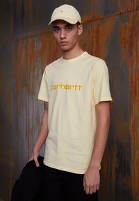 T-shirt de algodão amarelo com um logo central em laranja "carhartt.", combinado com uma boné amarelo correspondente. O modelo usa calças pretas, em frente a um fundo texturizado.