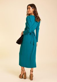 Robe midi turquoise avec manches longues, détail de taille noué, texture lisse et coupe droite, associée à des sandales à plateforme et une pochette noire.