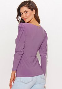 Fioletowa bluzka z długim rękawem z bufkami, dopasowany fason i okrągły dekolt. Posiada mały złoty guzik akcentujący z tyłu.