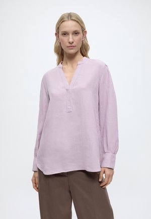 Moteris vilki šviesiai violetinę ilgomis rankovėmis palaidinę su V formos iškirpte ir rudus plačiomis kojomis kelnes, stovėdama baltame fone.