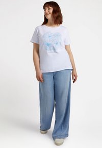 T-shirt bianca a maniche corte con un motivo floreale blu in linea, abbinata a jeans a gamba larga azzurri e sneakers beige.