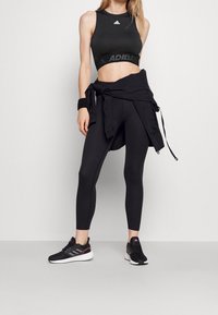 Top deportivo negro corto con logo, leggings negros a juego y zapatillas negras con acentos rosas. Presenta una chaqueta negra atada y una pulsera.
