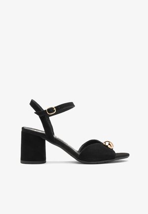 Czarne zamszowe sandały na obcasie z otwartym toe, z szerokim obcasem w kształcie bloku, regulowanym paskiem wokół kostki i ozdobnym złotym okrągłym akcentem na górze.