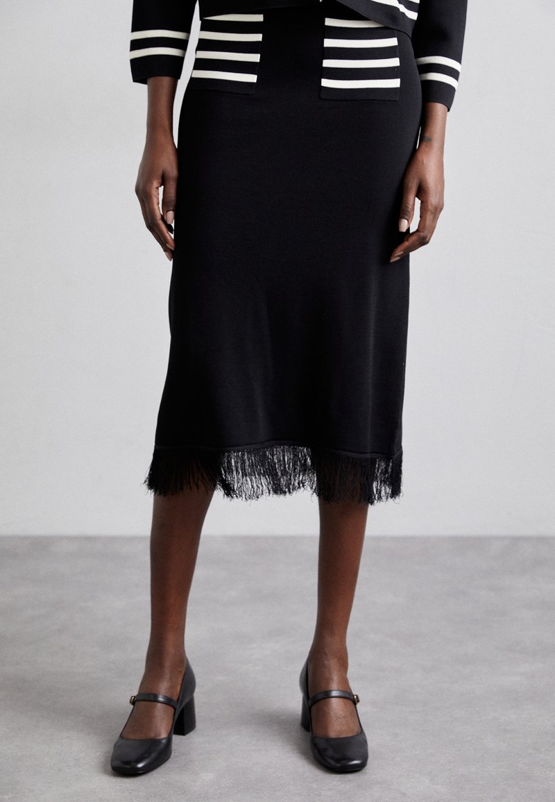 Jupe en tricot noire avec un ourlet à franges, présentant une silhouette ajustée. Associée à des chaussures noires à talons blocs avec une sangle sur le pied.