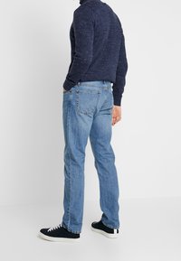 Blå denimjeans med rak passform, som har två bakfickor och synlig söm, i kombination med en mörk texturerad tröja och svarta sneakers.