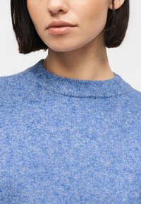 Γυναίκα με κοντά σκούρα μαλλιά που φοράει ελαφρύ γαλάζιο πλεκτό πουλόβερ με ριμπ πλεκτό λαιμόκοψη, μπροστά σε απλό λευκό φόντο.