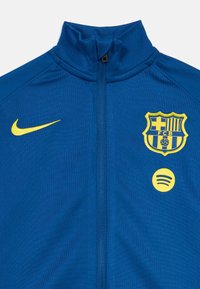 Μπλε ζακέτα με φερμουάρ κατασκευασμένη από πολυεστέρα, με κίτρινο λογότυπο Nike, έμβλημα της Μπαρτσελόνα και λογότυπο Spotify στο μπροστινό μέρος. Λείο υλικό.