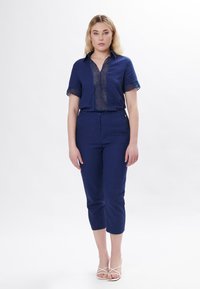 Navy linnen overhemd met korte mouwen, voorzien van verticale uitsparingen. Gecombineerd met strakke navy capri broek. Model draagt witte sandalen met bandjes.