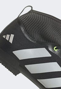 Chaussure de soccer noire avec une tige en tissu texturé, dotée de trois rayures argentées diagonales et d'un accent vert au niveau de la boucle du col. Semelle en caoutchouc.
