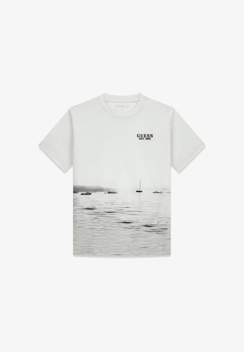 T-shirt en coton gris clair avec une impression photographique de bateaux sur l'eau. Comprend le texte "GUESS EST. 1981" en noir sur la poitrine. Manches courtes.