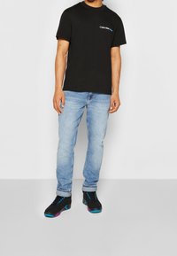 Černé bavlněné tričko s logem, světle modré džíny a černé tenisky s modrými a růžovými akcenty, stojící na jednoduchém pozadí.