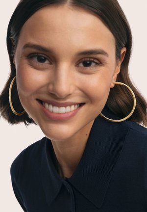 Primer plano de una persona con cabello corto y oscuro que lleva grandes pendientes de aro dorados con textura. Viste una camiseta de cuello azul marino y sonríe.