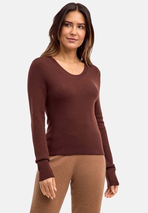 Donna con lunghi capelli scuri che indossa un maglione aderente marrone a maniche lunghe e pantaloni marrone chiaro, in piedi con uno sfondo grigio neutro.
