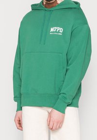 Sudadera verde hecha de algodón suave, con un bolsillo tipo canguro, capucha con cordón y un logotipo en texto blanco en el pecho. Presenta puños y dobladillo acanalados.